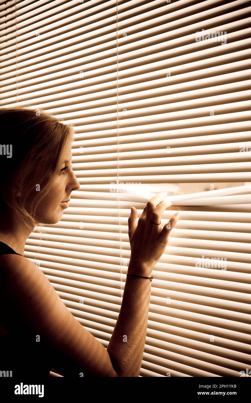 Eine blonde Frau, die neben einem Fenster mit Fensterläden steht und nach draußen schaut Stockfoto