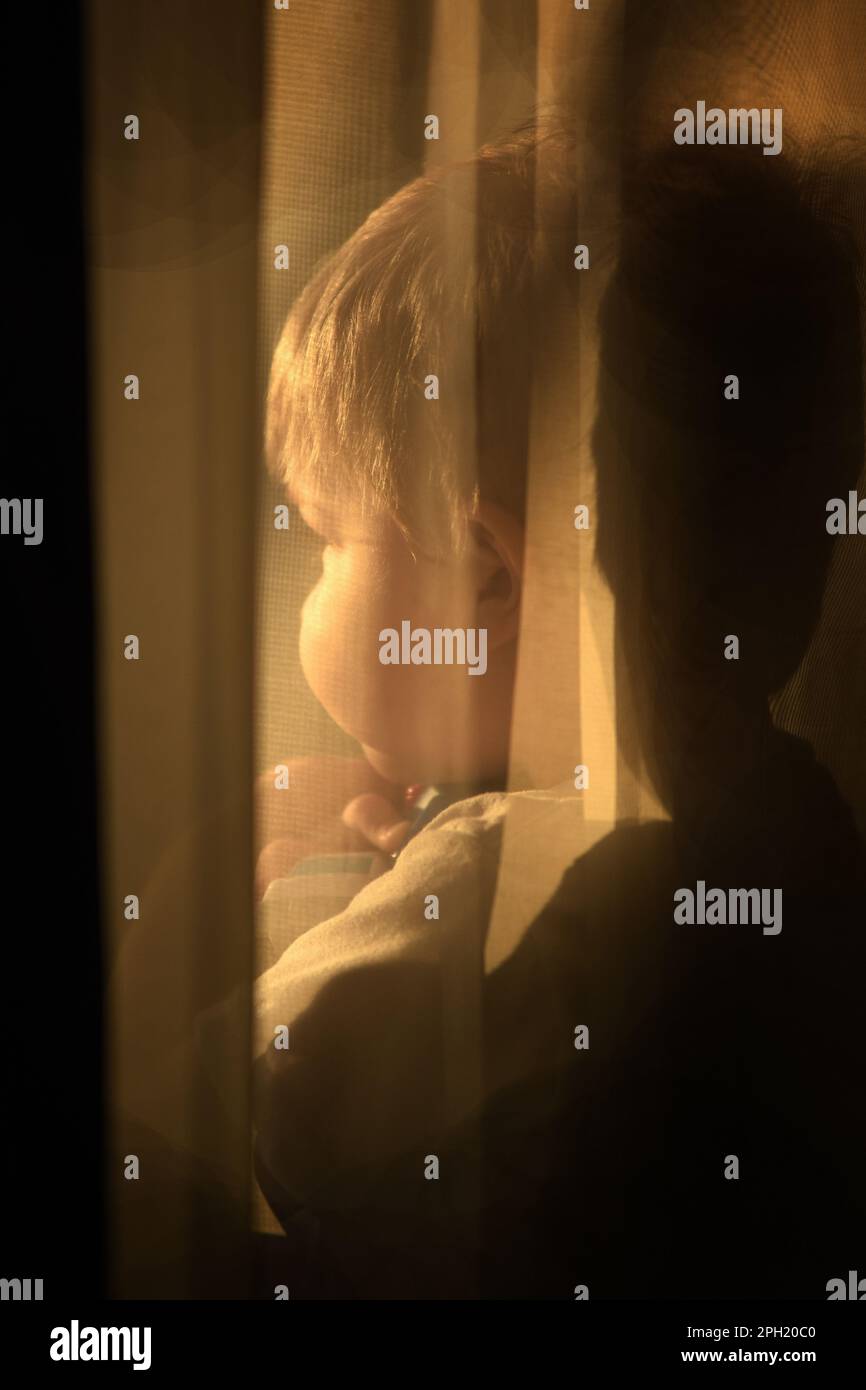 Ein 2-jähriger Junge, der hinter durchsichtigen Vorhängen aus dem Fenster schaut. Stockfoto
