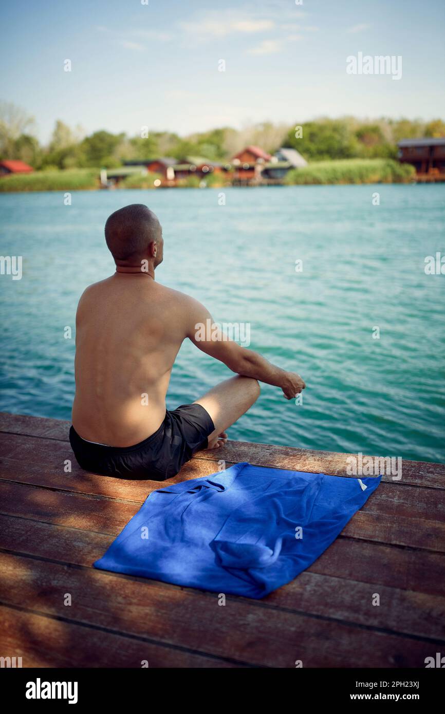 Ein Mann, der morgens auf dem Wasser meditiert. Junger Mann, der auf einem hölzernen Steg am Meer sitzt und spirituelle Übungen macht. Entspannung, Disziplin, Lifestyle-Konzept. Stockfoto