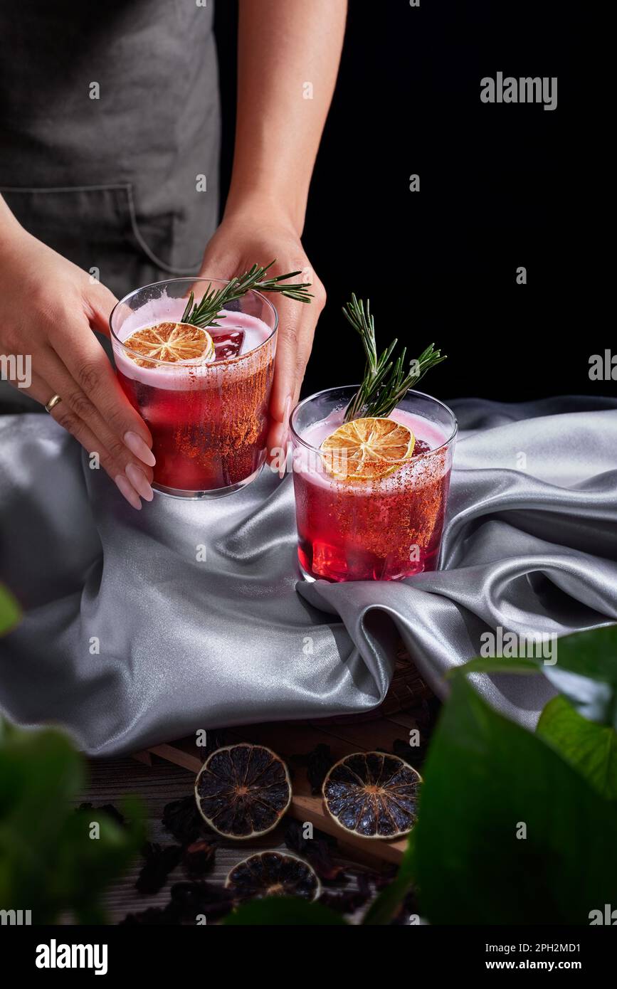 Roter Getränk-Cocktail aus Rum und Mezcal mit Limetten- und Kirschsaft garniert mit getrockneten Früchten und grünem Rosmarin, platziert von zwei weiblichen Händen, Pflanzen Stockfoto