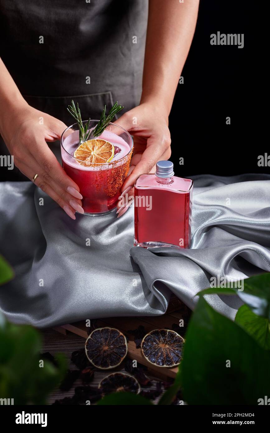Ein Glas Getränke mit einem Cocktail aus rotem Rum und einem Mezcal-Getränk mit Limetten- und Kirschsaft garniert mit getrockneten Früchten und grünem Rosmarin, platziert von zwei Weibchen Stockfoto