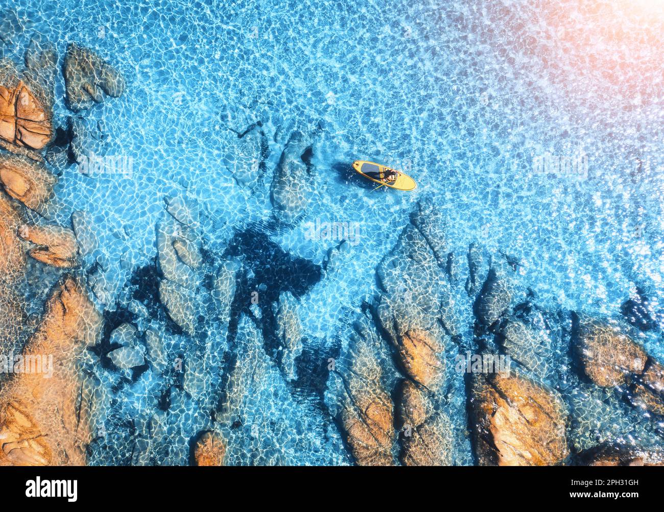 Luftaufnahme des Mannes auf dem schwimmenden Sup Board auf blauem Meer, felsiger Strand Stockfoto