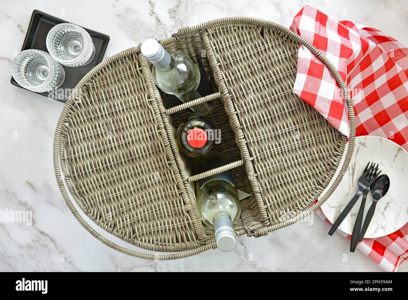Picknickkorb aus Korb mit Weinfach, Weinflaschen, Weingläsern aus Kunststoff und Besteck mit rot-weiß kariertem Tuch auf Marmorhintergrund im fl Stockfoto