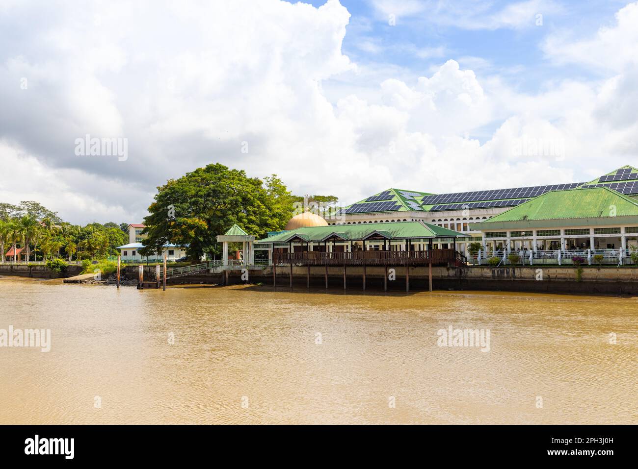 Gebäude und Bäume entlang des Flusses Sungai Temburong im Bezirk Temburong in Brunei Darussalam, mit blauem Himmel und gelbem Wasser Stockfoto