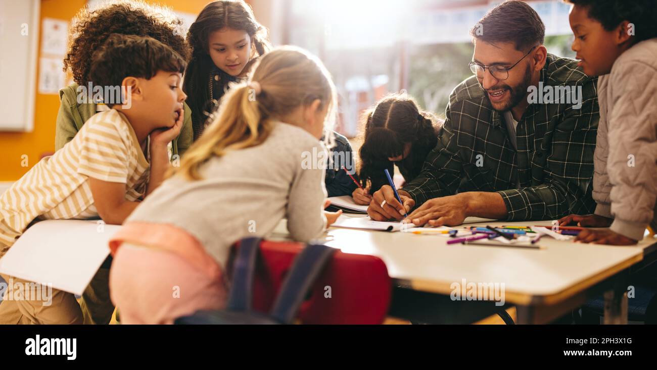 Eine Gruppe von Grundschulkindern steht in einem Klassenzimmer um einen Tisch und folgt aufmerksam einer Lektion, die ihr Lehrer erteilt. Junge Studenten enga Stockfoto