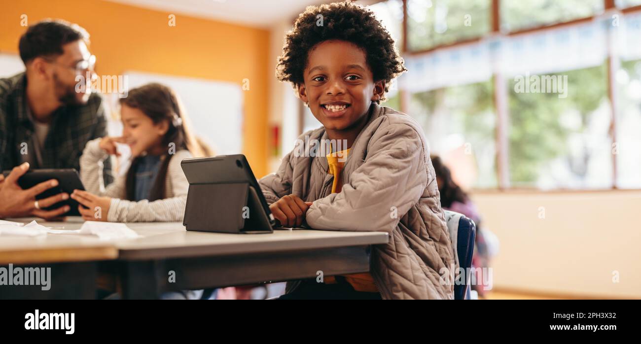 Ein Junge lächelt in die Kamera, während er in einem Klassenzimmer mit einem digitalen Tablet sitzt. Ein Kind aus der Grundschule liest ein Buch in der Schule. Kleines Kind entwickelt seine Stockfoto