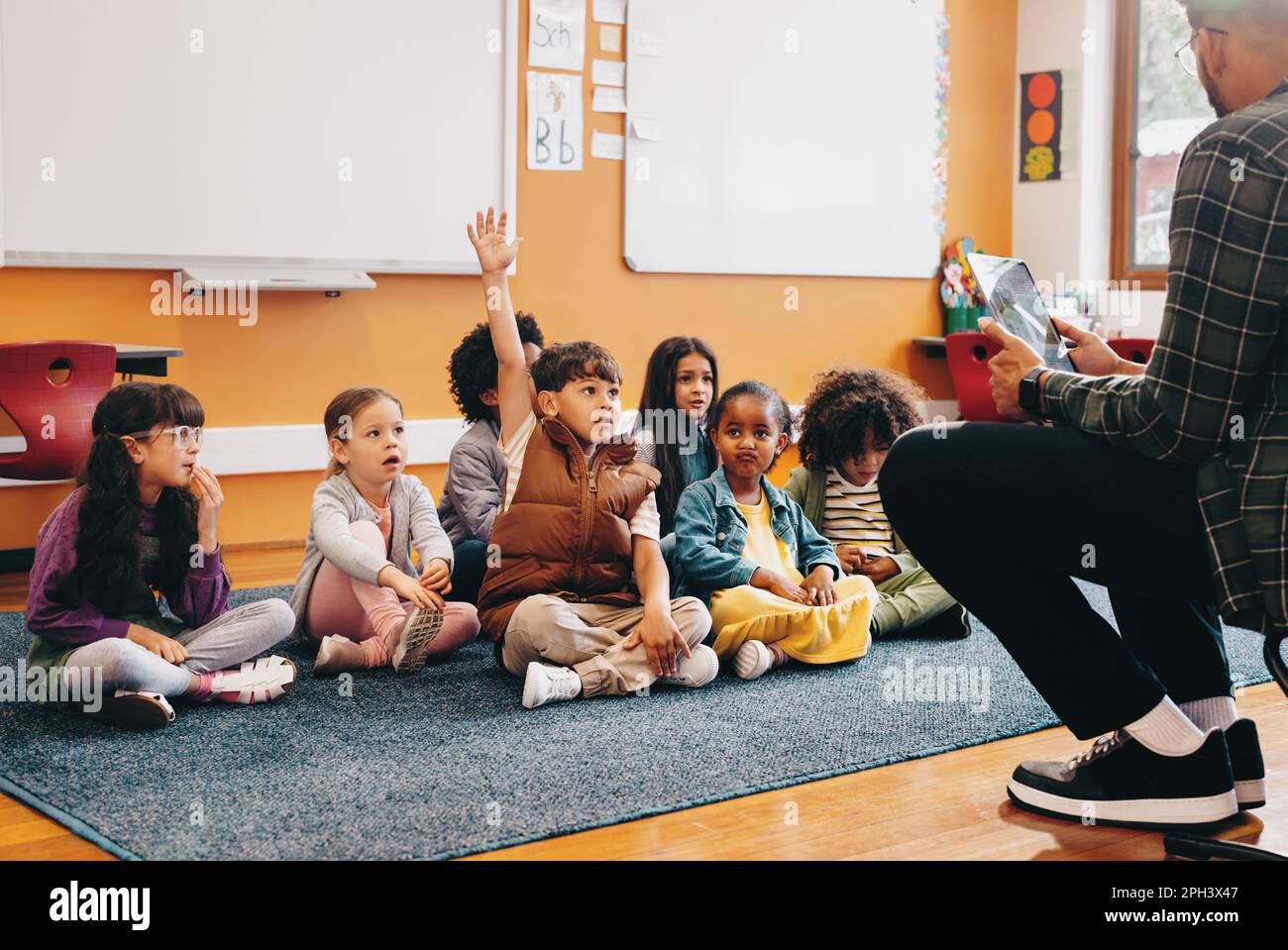 Der Junge hebt die Hand, um eine Frage in einem Klassenzimmer zu beantworten; er sitzt mit anderen Kindern auf dem Boden und der Lehrer sitzt vor der Klasse. Stockfoto