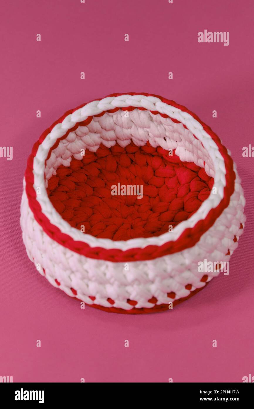 Häkelkorb auf pinkfarbenem Hintergrund. Rot-weiße runde Box für Kleinigkeiten aus gestricktem Garn, handgefertigter Kunst. Draufsicht. Stockfoto