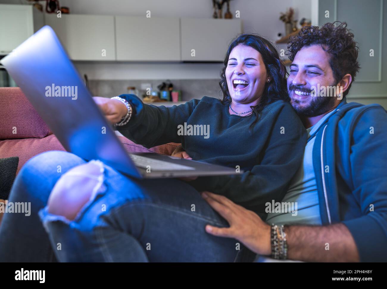 Ein junges Paar kuschelt auf einer gemütlichen Couch, während es auf der Suche nach preisgünstigen Reiseangeboten mit einem Laptop ist. Das lächelnde Duo wirkt entspannt und zufrieden Stockfoto