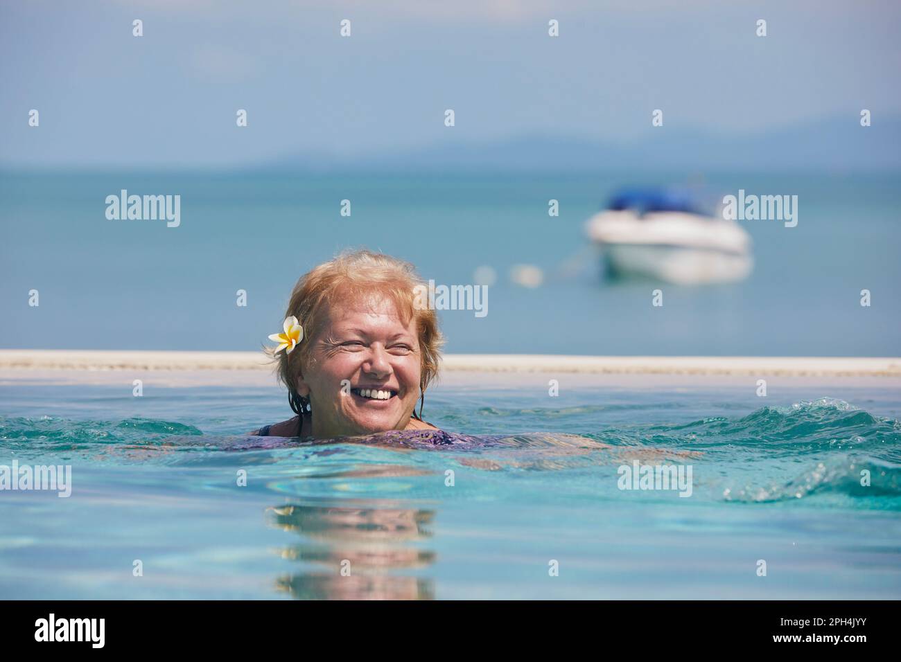 Aktive, lachende Seniorin im Pool gegen das Meer. Die Frau schwimmt an sonnigen Tagen. Stockfoto