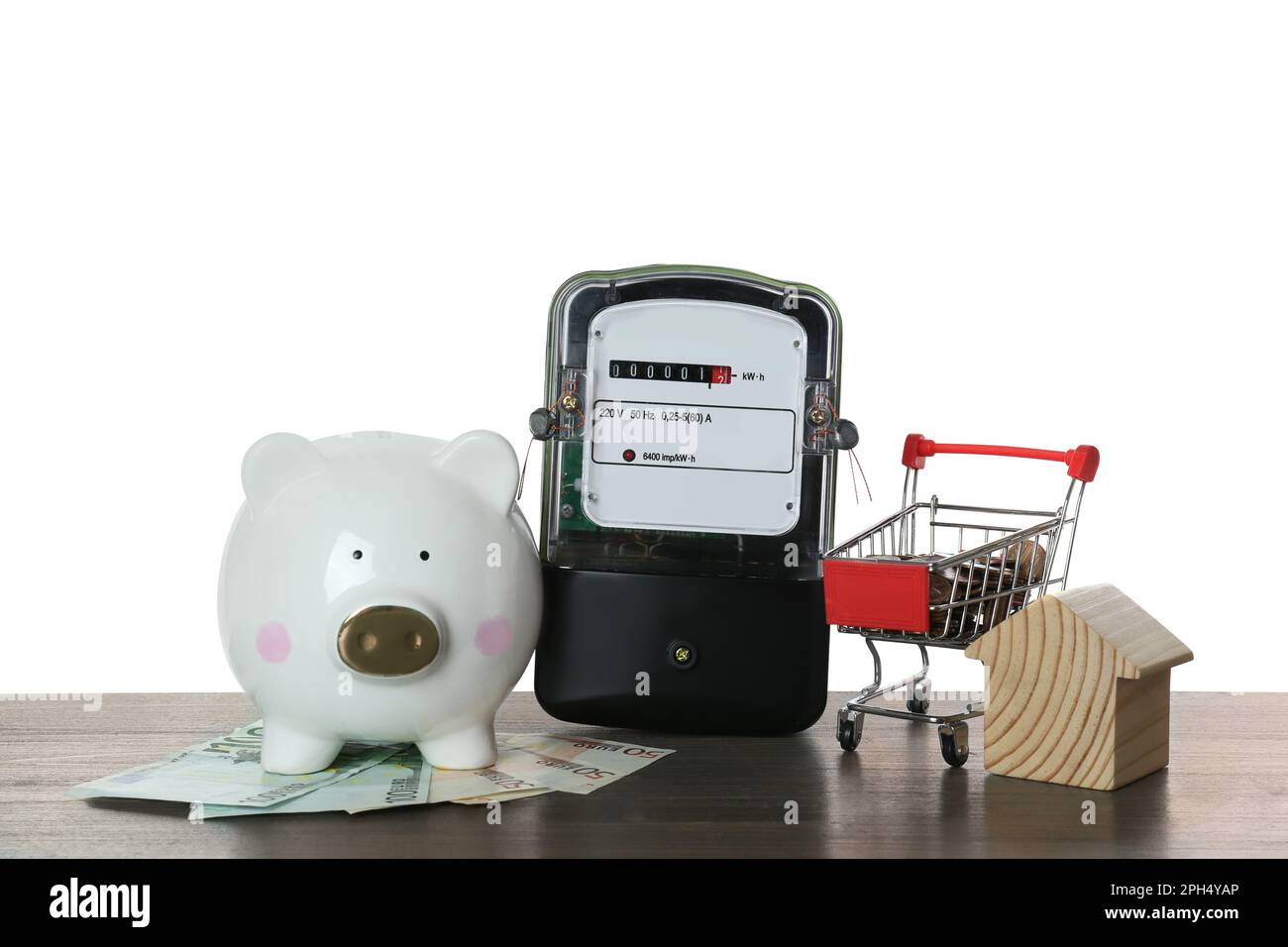 Stromzähler, Hausmodell, Sparschwein und Einkaufswagen mit Geld auf Holztisch vor weißem Hintergrund Stockfoto