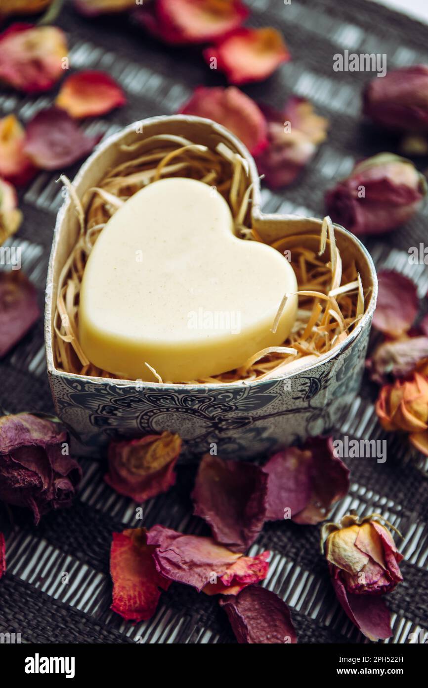 Kräftige, cremefarbene Riegel in einer herzförmigen Geschenkbox auf schwarzem Hintergrund, dekoriert mit trockenen Rosenblütenblättern im Innenbereich. Stockfoto
