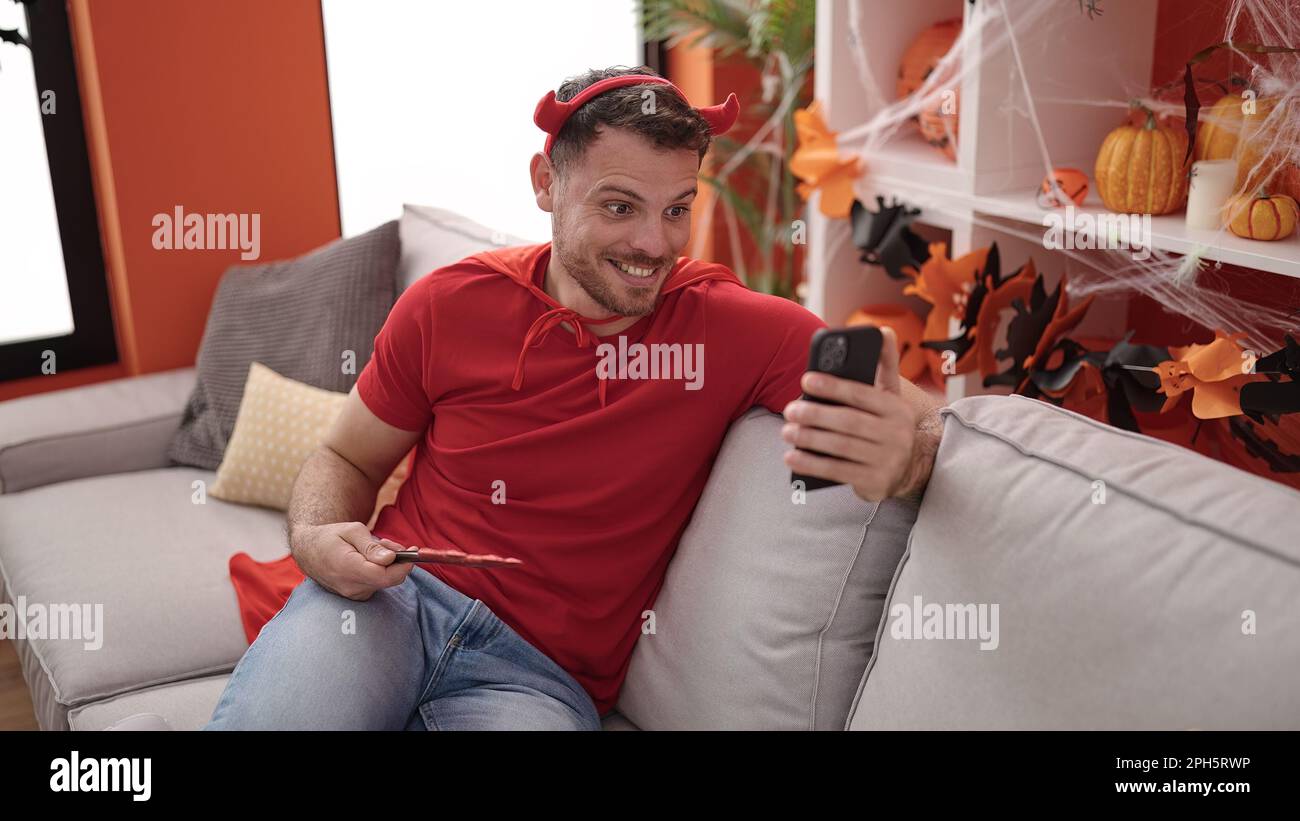 Junger Kaukasier in Teufelskostüm, der auf dem Sofa sitzt und zu Hause Videoanrufe macht Stockfoto