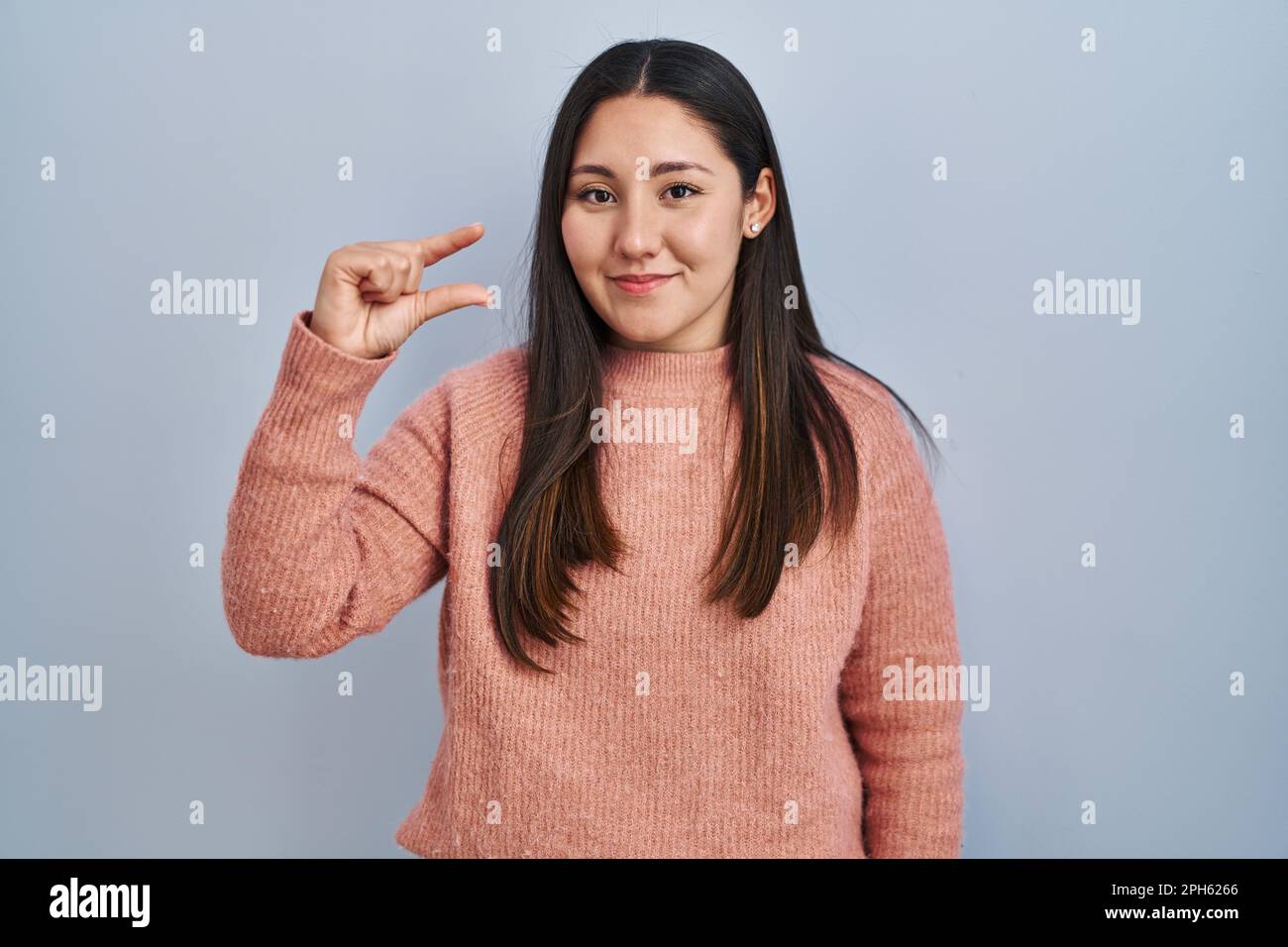 Junge lateinische Frau, die über blauem Hintergrund lächelt und selbstbewusst gestikelt, mit der Hand kleine Zeichen tut, mit den Fingern schaut und der Kamera. M Stockfoto