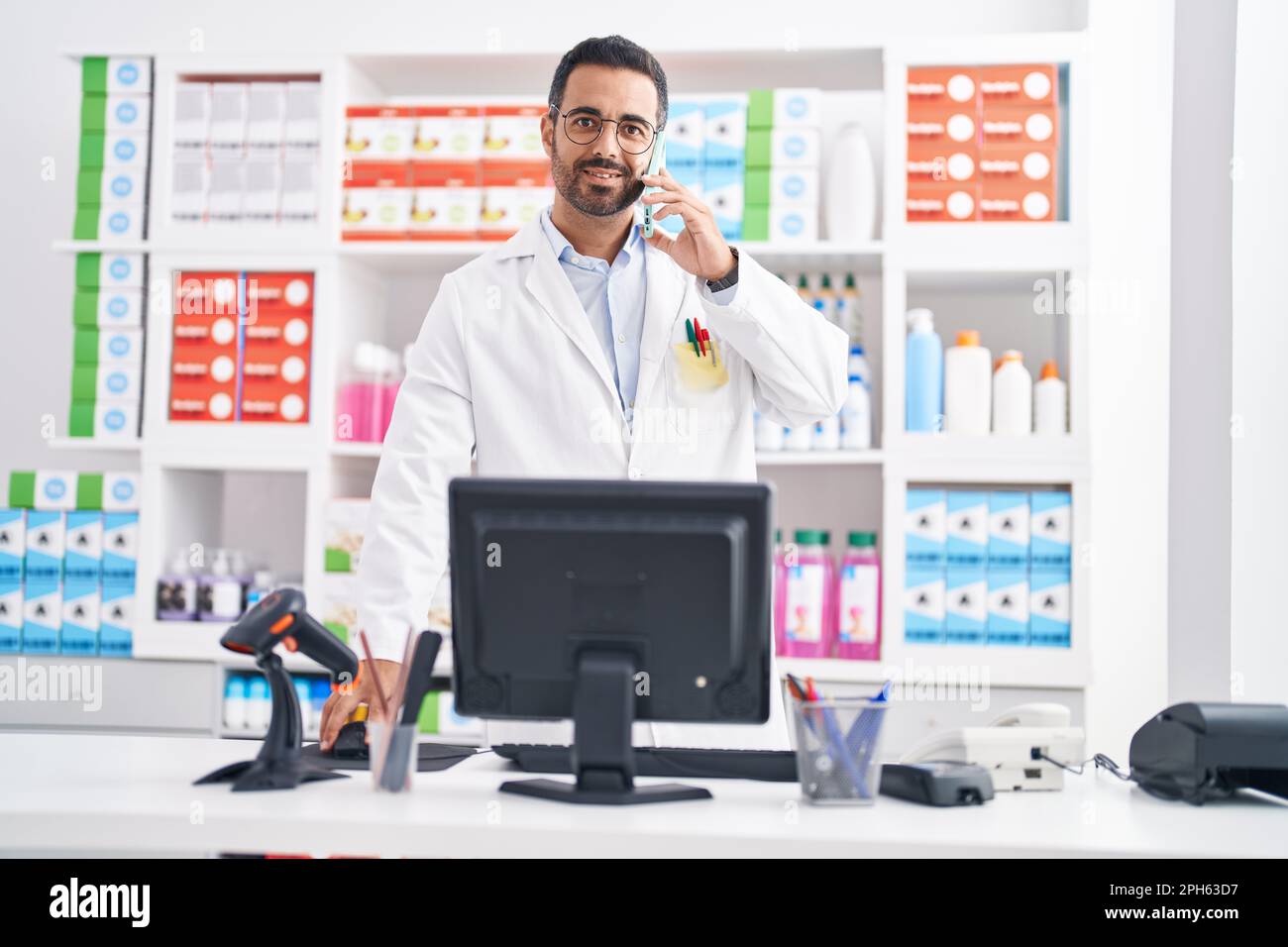 Junger hispanic-Mann Apotheker im Gespräch auf Smartphone mit Computer in der Apotheke Stockfoto