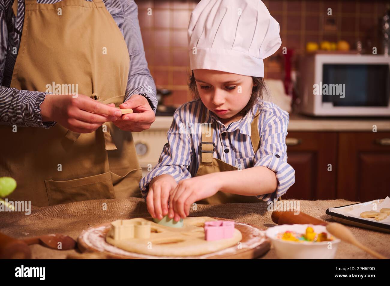 Ein Kind mit Kochmütze bereitet Osterkuchen zu und schneidet Teig unter Anleitung ihrer mutter mit einem Ausstecher Stockfoto