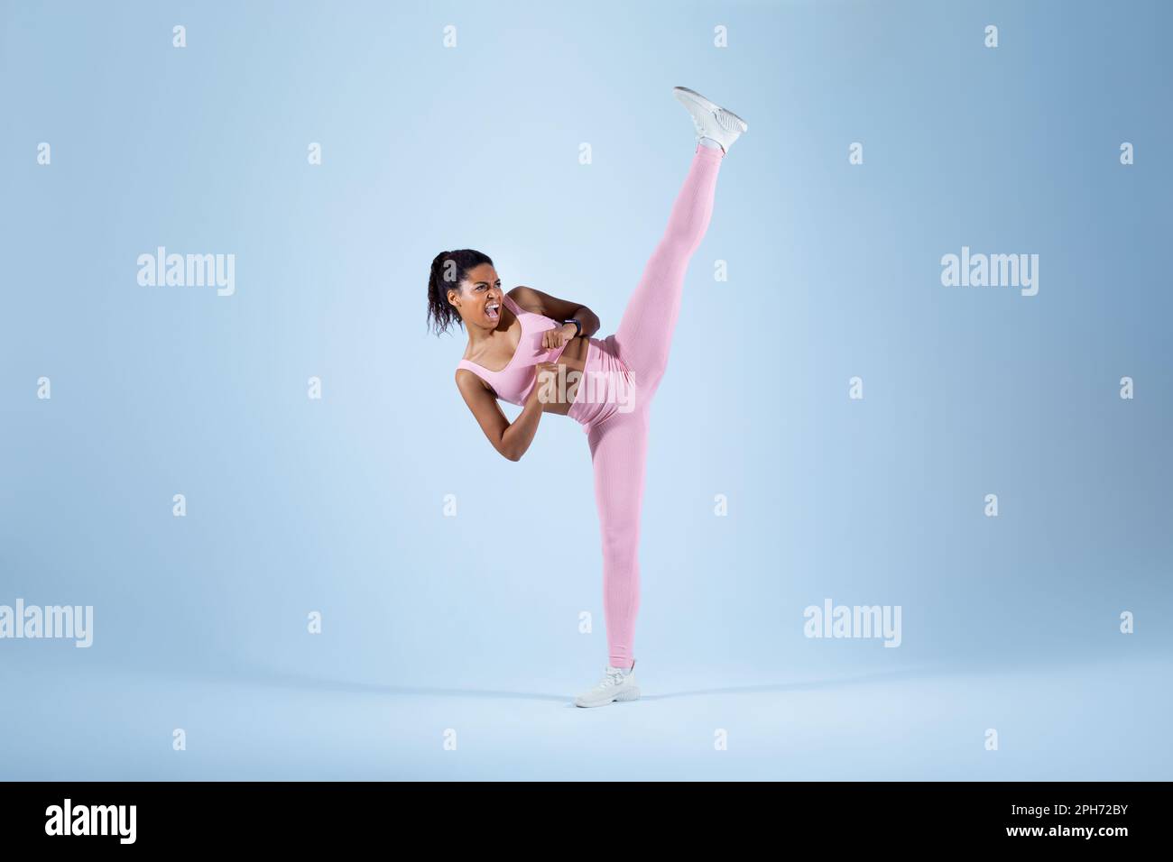 Eine sportliche schwarze Frau, die Karate-Side-Kick macht, die Fäuste zusammendrückt und das Bein hebt, und über einem blauen Neon-Studiohintergrund steht Stockfoto