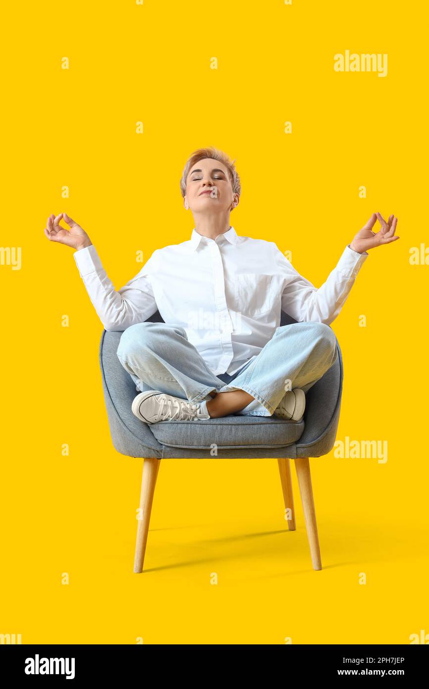 Meditierende reife Frau, die im Sessel auf gelbem Hintergrund sitzt Stockfoto