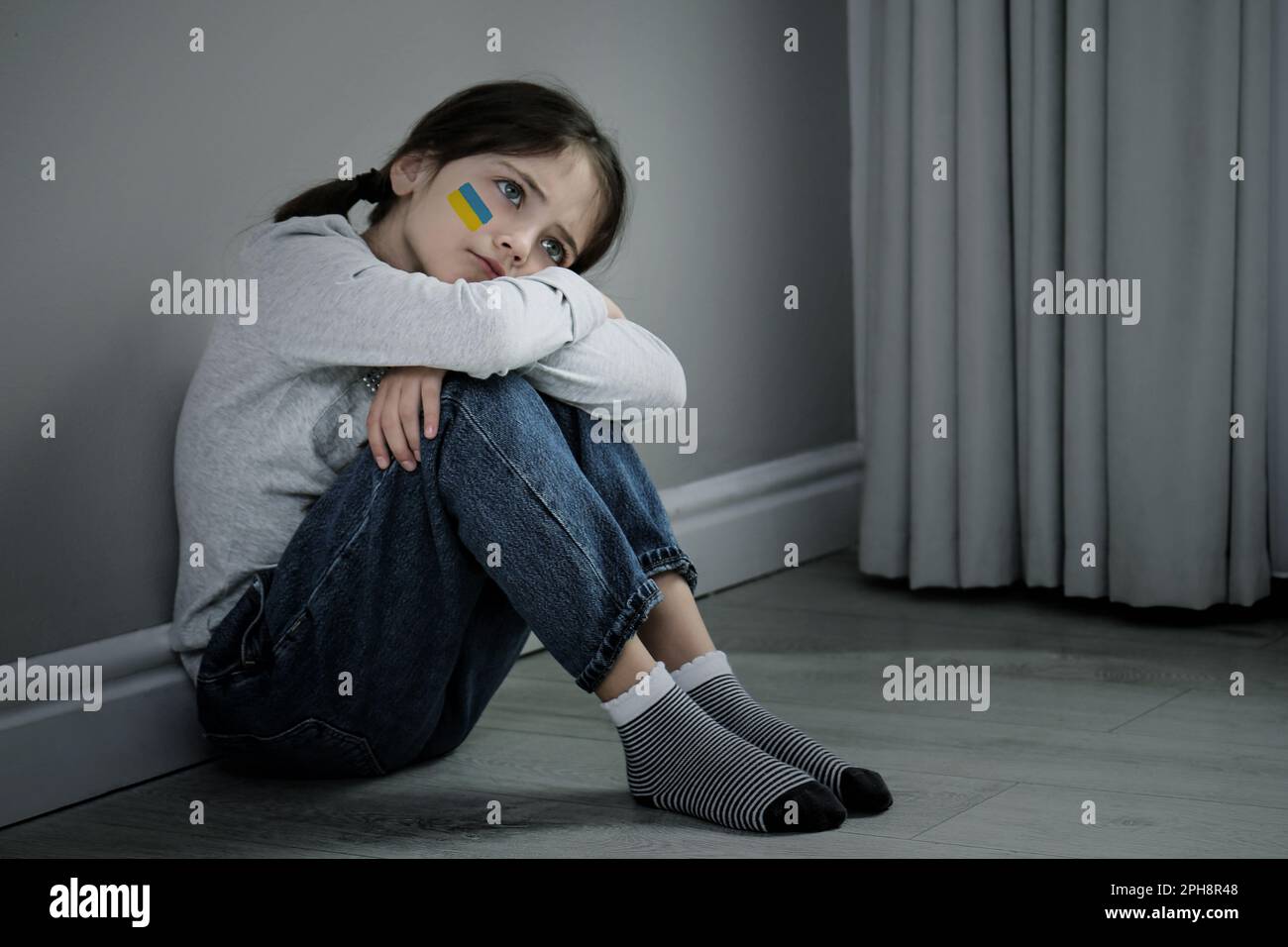 Verärgertes kleines Mädchen mit dem Bild der ukrainischen Flagge auf der Wange, das drinnen auf dem Boden sitzt. Beenden Sie den Krieg in der Ukraine Stockfoto