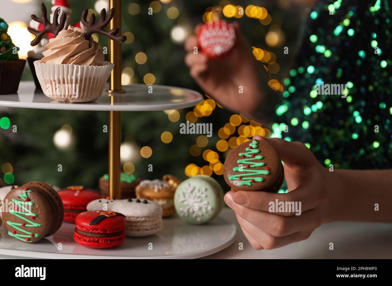 Eine Frau, die Weihnachtsmakaron aus dem Stand nimmt, gegen verschwommene festliche Lichter, Nahaufnahme Stockfoto