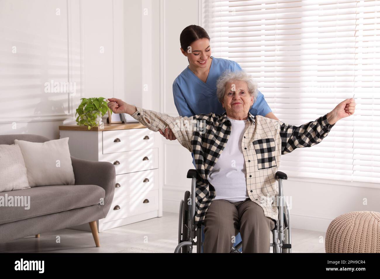 Seniorin im Rollstuhl, die körperliche Bewegung macht, und junge Pflegekraft, die ihr drinnen hilft. Häuslicher Gesundheitsdienst Stockfoto