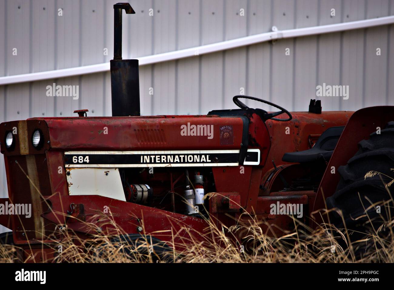 Ein alter rostbedeckter roter Traktor, der sich in einem Zustand der Reparatur befindet, steht auf einem Feld mit hohem Unkraut Stockfoto