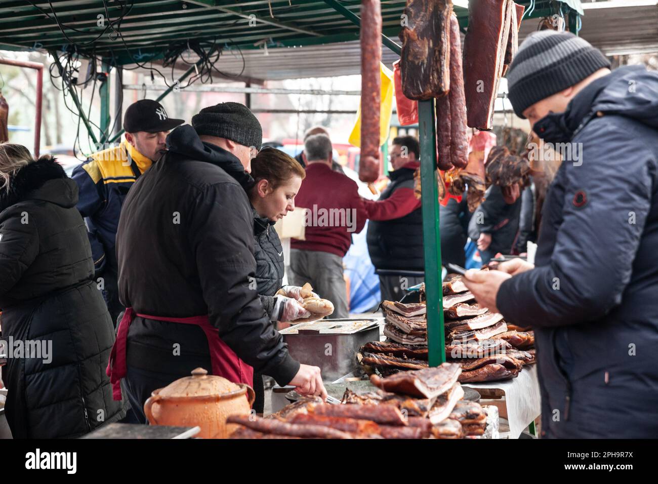 Bild eines Stalls des Kacarevo Marktes in Serbien, bei dem die Kunden das aufgehängte und trocknende Fleisch, wie Schinken und Würstchen, verkosten. Stockfoto