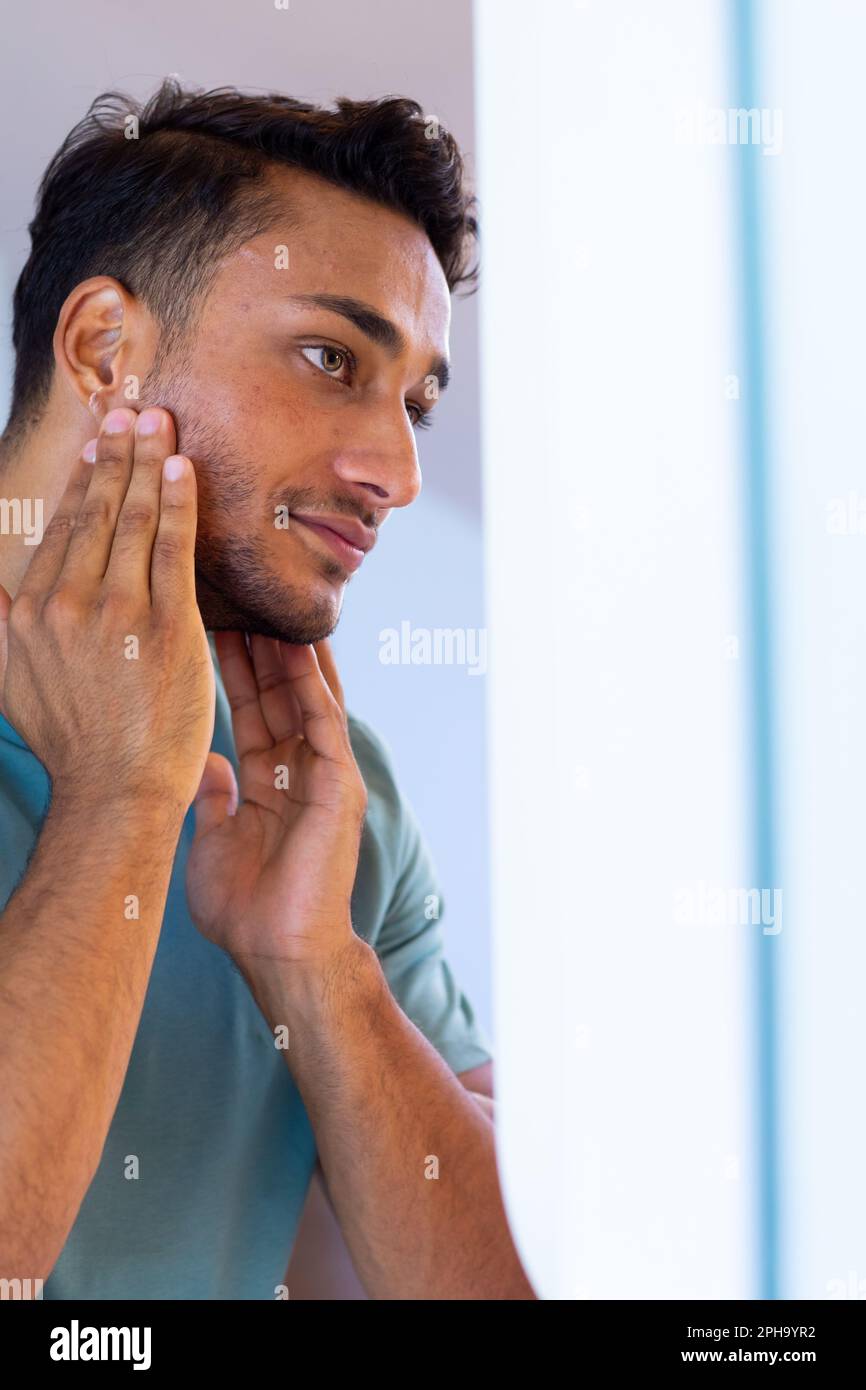 Lächelnder, birassistischer Mann, der sich im Badezimmerspiegel ansieht und Gesichtscreme aufträgt Stockfoto