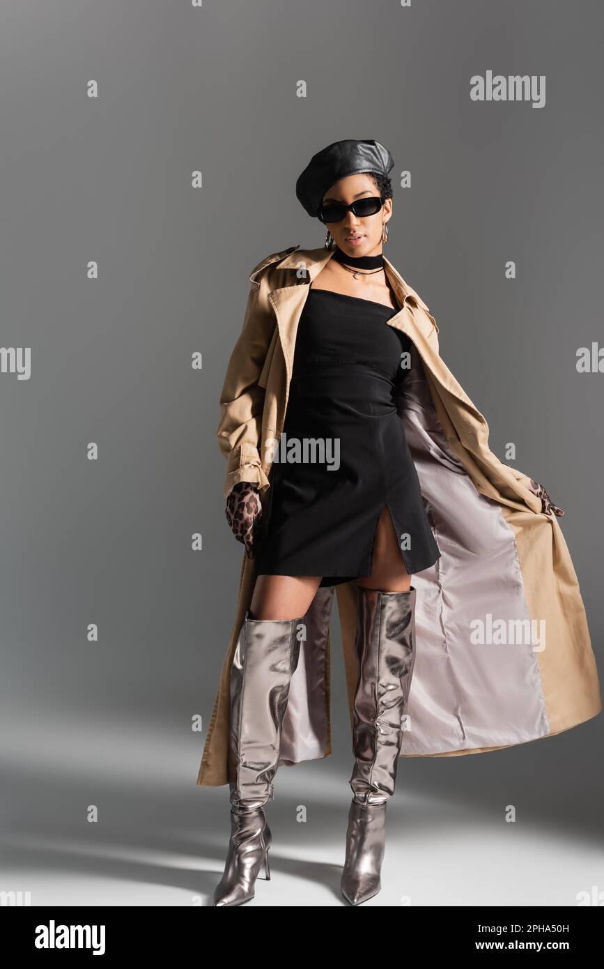 Trendige afroamerikanische Frau in Trenchcoat und glänzenden Stiefeln auf grauem Hintergrund, Stockbild Stockfoto