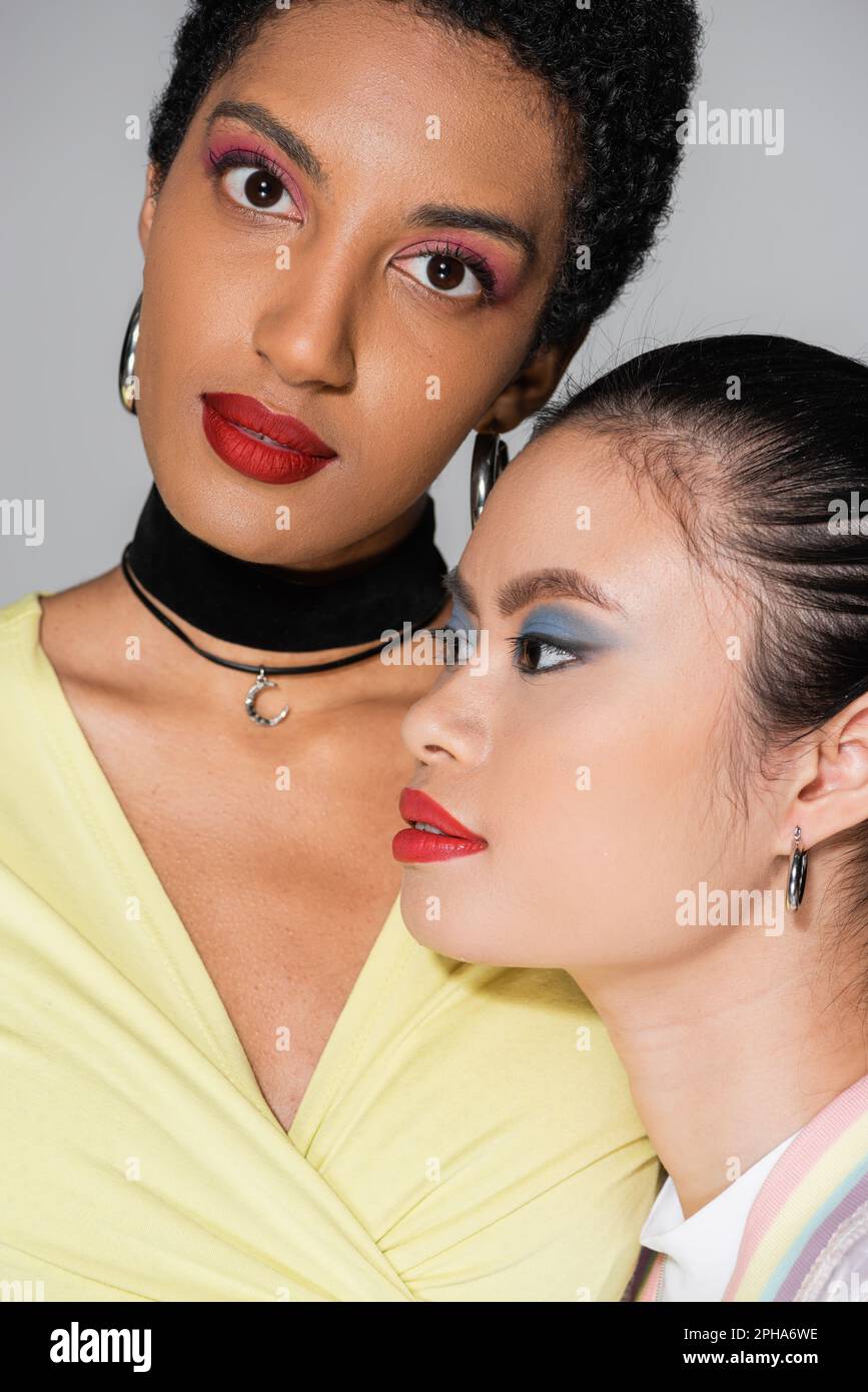 Porträt einer kurzhaarigen afroamerikanischen Frau, die in der Nähe eines asiatischen Freundes in die Kamera schaut, mit roten Lippen isoliert auf grauem Stockbild Stockfoto