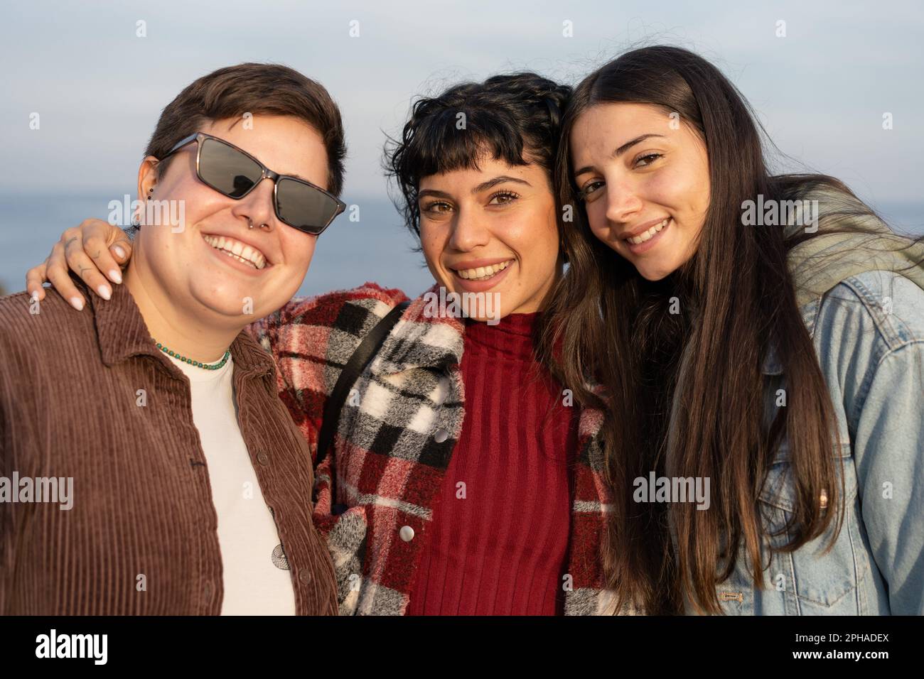 Drei verschiedene weibliche Freunde umarmen und lächeln am Meer - eine nicht binäre Übergröße mit Sonnenbrille, kurzes Haar, Brünette mit welligen Haaren und eine andere Stockfoto