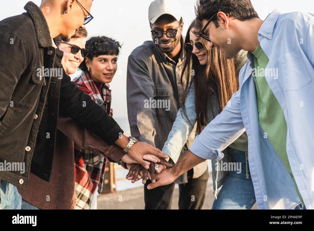 Eine vielfältige Gruppe von Freunden, die sich mit Afrikanern, Kaukasiern und einer nicht-binären Person mit kurzen Haaren zusammenschließen, Hand in Hand. Sie stehen mit T am Meer Stockfoto