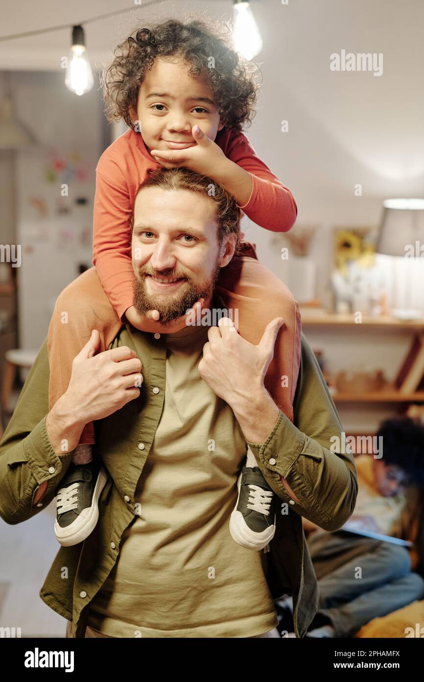 Glücklicher junger Mann, der im Wohnzimmer in die Kamera schaut und seinen niedlichen kleinen Sohn auf den Schultern hält, während er Spaß hat Stockfoto