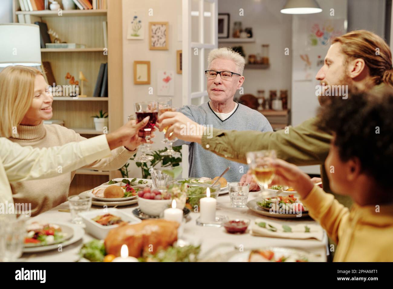 Eine Gruppe von Familiensäugern, die mit Gläsern hausgemachtem Wein über dem Tisch klimpern, serviert mit Speisen und dekoriert mit brennenden Kerzen Stockfoto