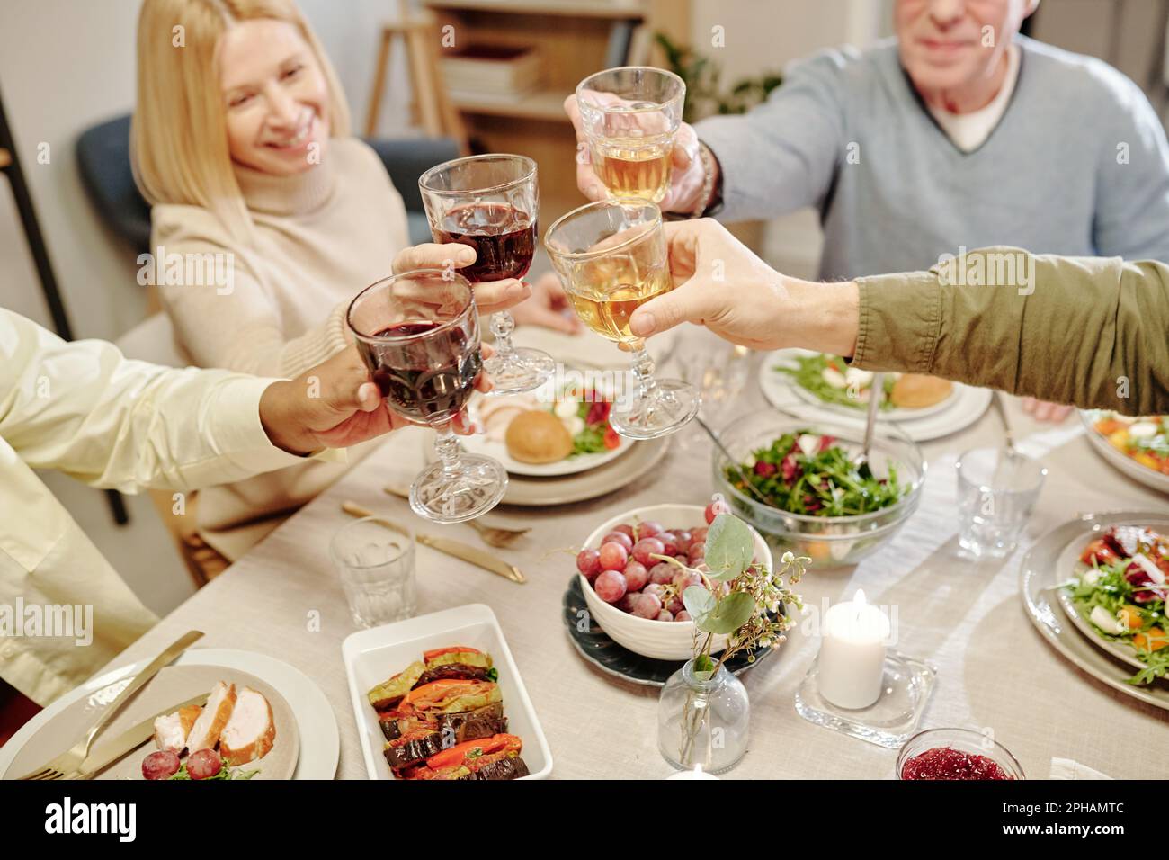 Interkulturelle Familienmitglieder stoßen mit einem hohen Winkel an den Händen mit Gläsern hausgemachtem Wein auf dem Tisch an, serviert mit leckeren Speisen Stockfoto