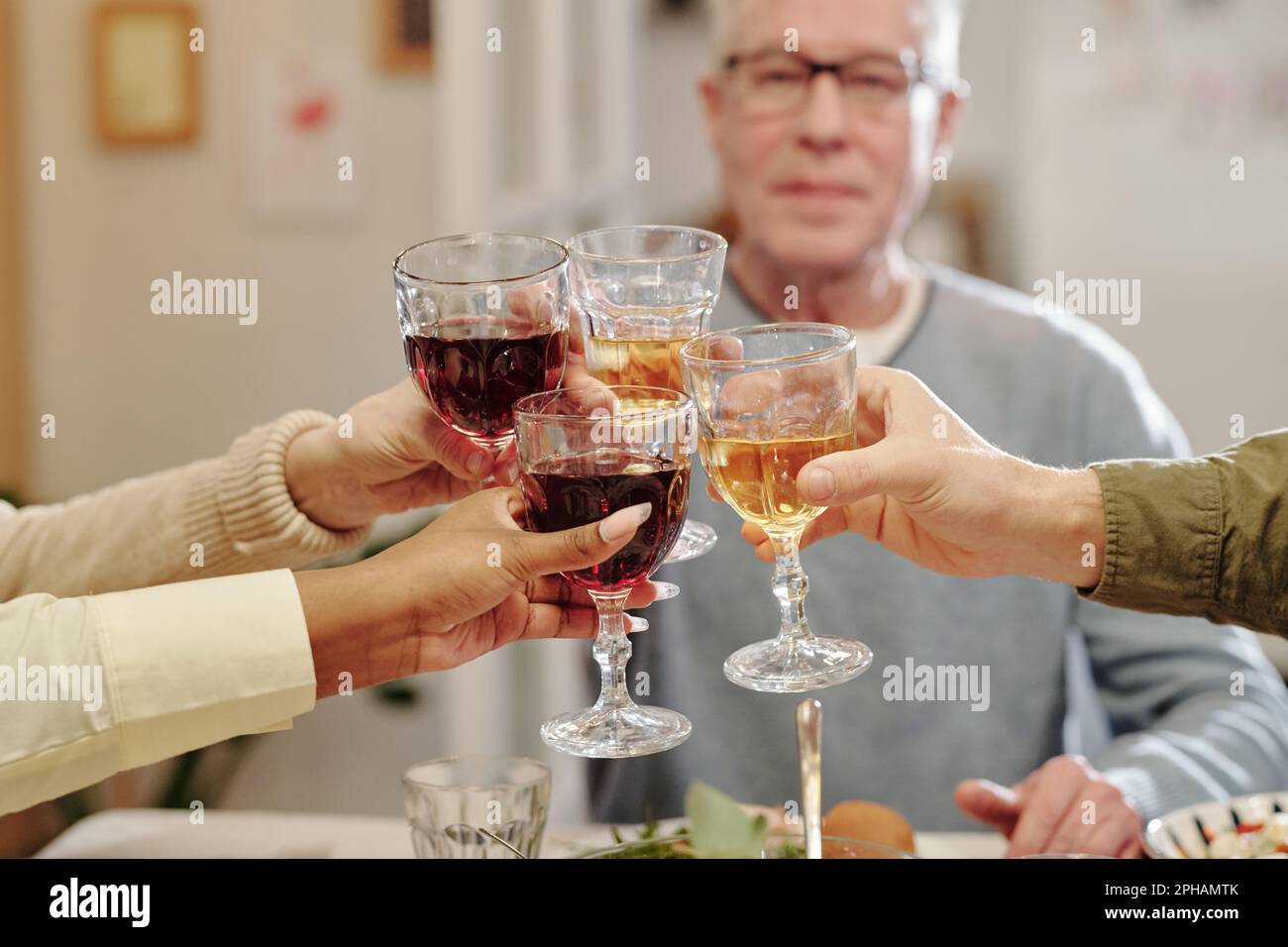 Nahaufnahme der Hände von Mitgliedern der interkulturellen Familie, die mit Gläsern hausgemachtem Wein über einem festlichen Tisch klimpern, während sie ein Lebensereignis feiern Stockfoto