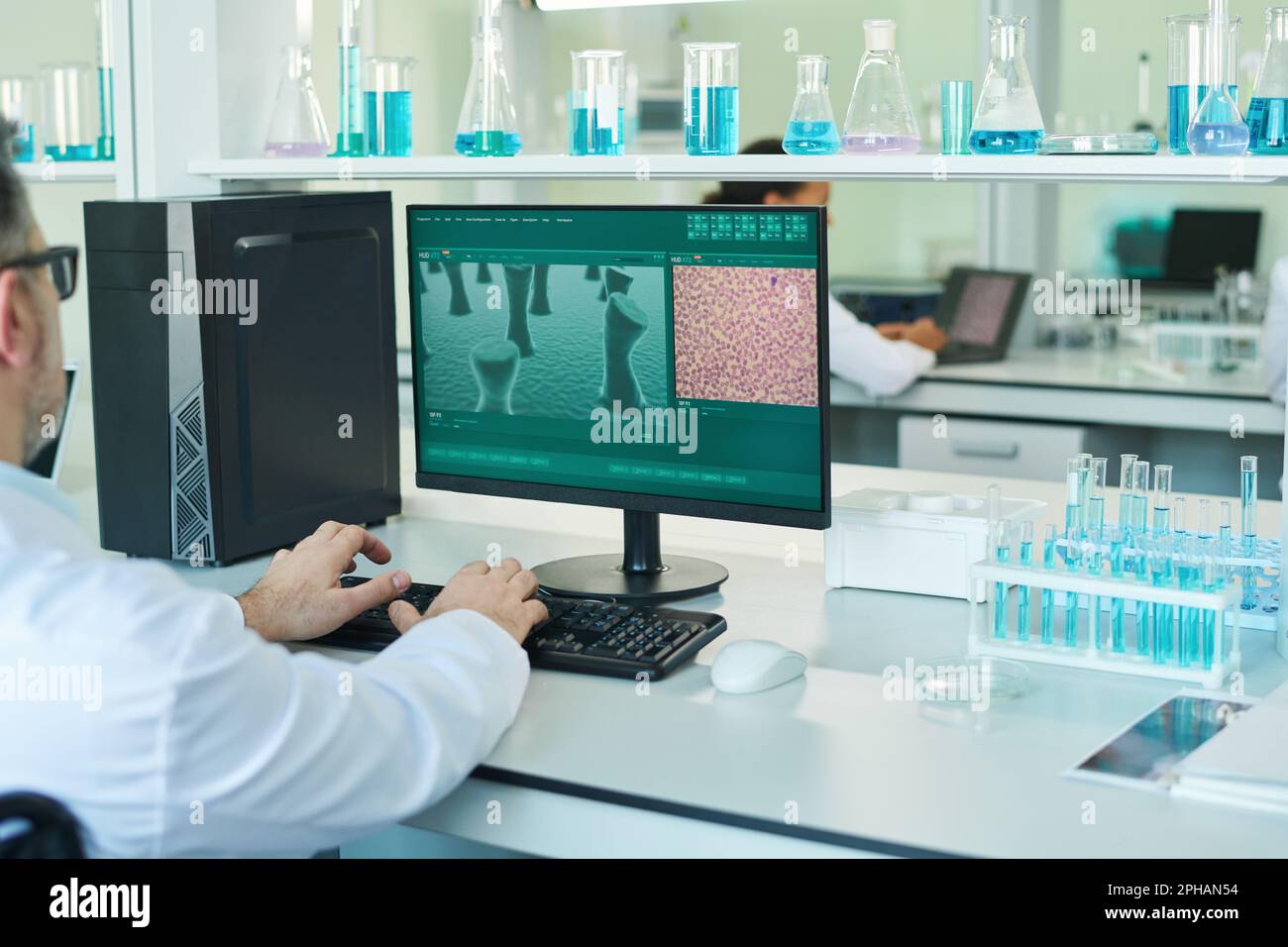 Arme eines erfahrenen Wissenschaftlers, der klinische Experimente durchführt, während er am Schreibtisch vor dem Computermonitor sitzt und einen Teil der Zelle auf dem Bildschirm anzeigt Stockfoto