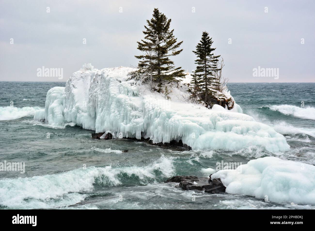 Die Wellen stürzen sich an einer Insel vorbei, die von Eis umgeben ist und von zwei harten immergrünen Bäumen gekrönt ist Stockfoto