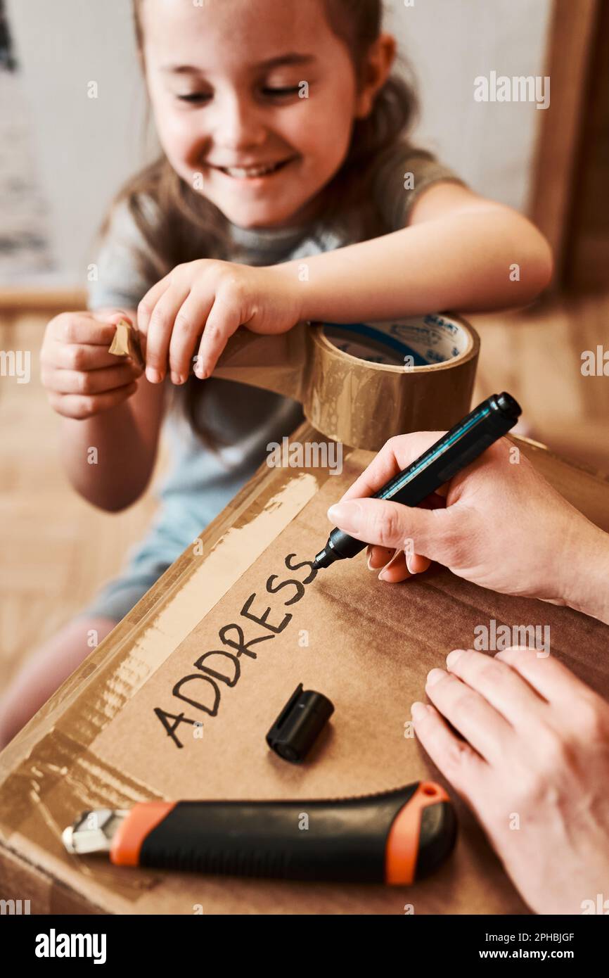 Frau, die eine Adresse auf ein Pappkartonpaket im Zimmer zu Hause schreibt. Kleines Mädchen