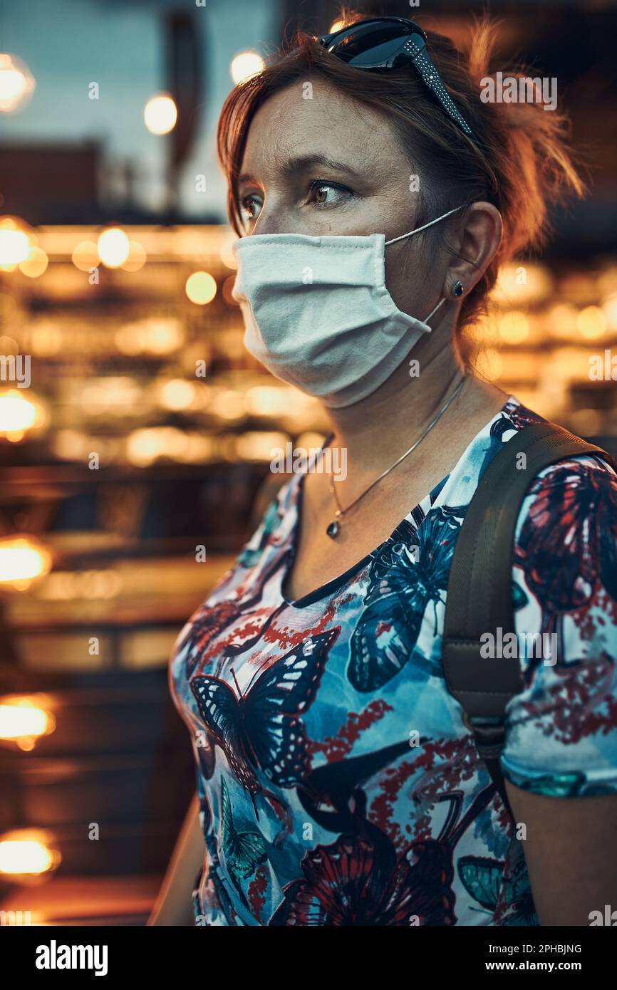 Junge Frau, die am Abend nach dem Einkaufen in der Innenstadt entlang einer Ladenfront geht, trägt die Gesichtsmaske, um Virusinfektion zu vermeiden und zu präven Stockfoto