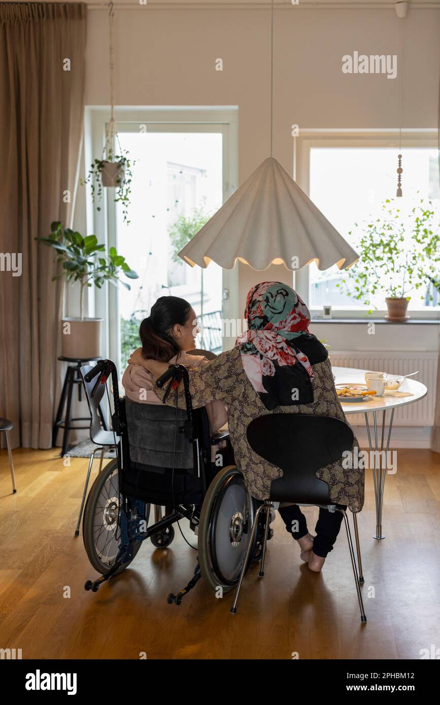 Eine Frau, die im Rollstuhl von der Mutter am Esstisch sitzt Stockfoto