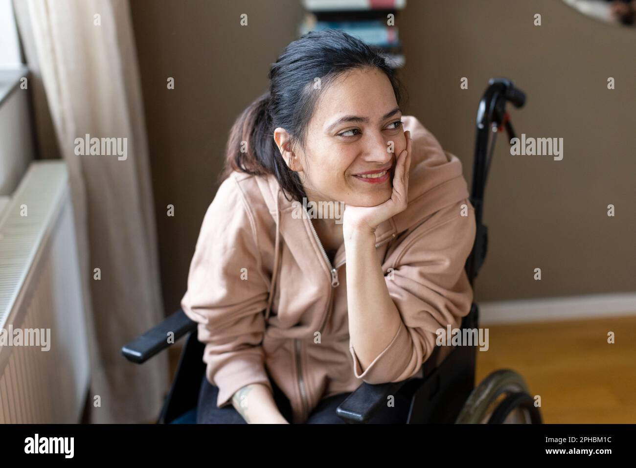 Lächelnde junge Frau mit Hand am Kinn, die zu Hause im Rollstuhl sitzt Stockfoto