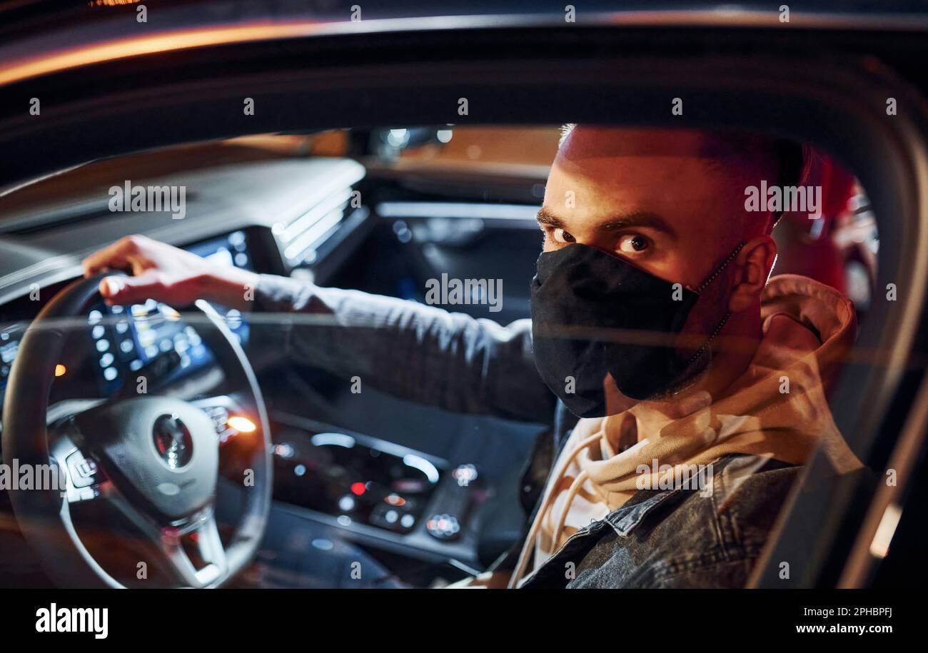 In schwarzer Schutzmaske. Ein gut aussehender, unrasierter Mann in modischer Kleidung, der sein Auto fährt Stockfoto