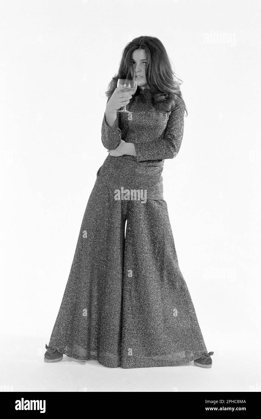 studio Portrait schlanke, hübsche Frau langes dunkles Haar, Hose mit sehr breiten Leuchtröhren, passend zum Oberteil, trinkt Cognac aus Glas mit weißem Hintergrund 1970er Stockfoto