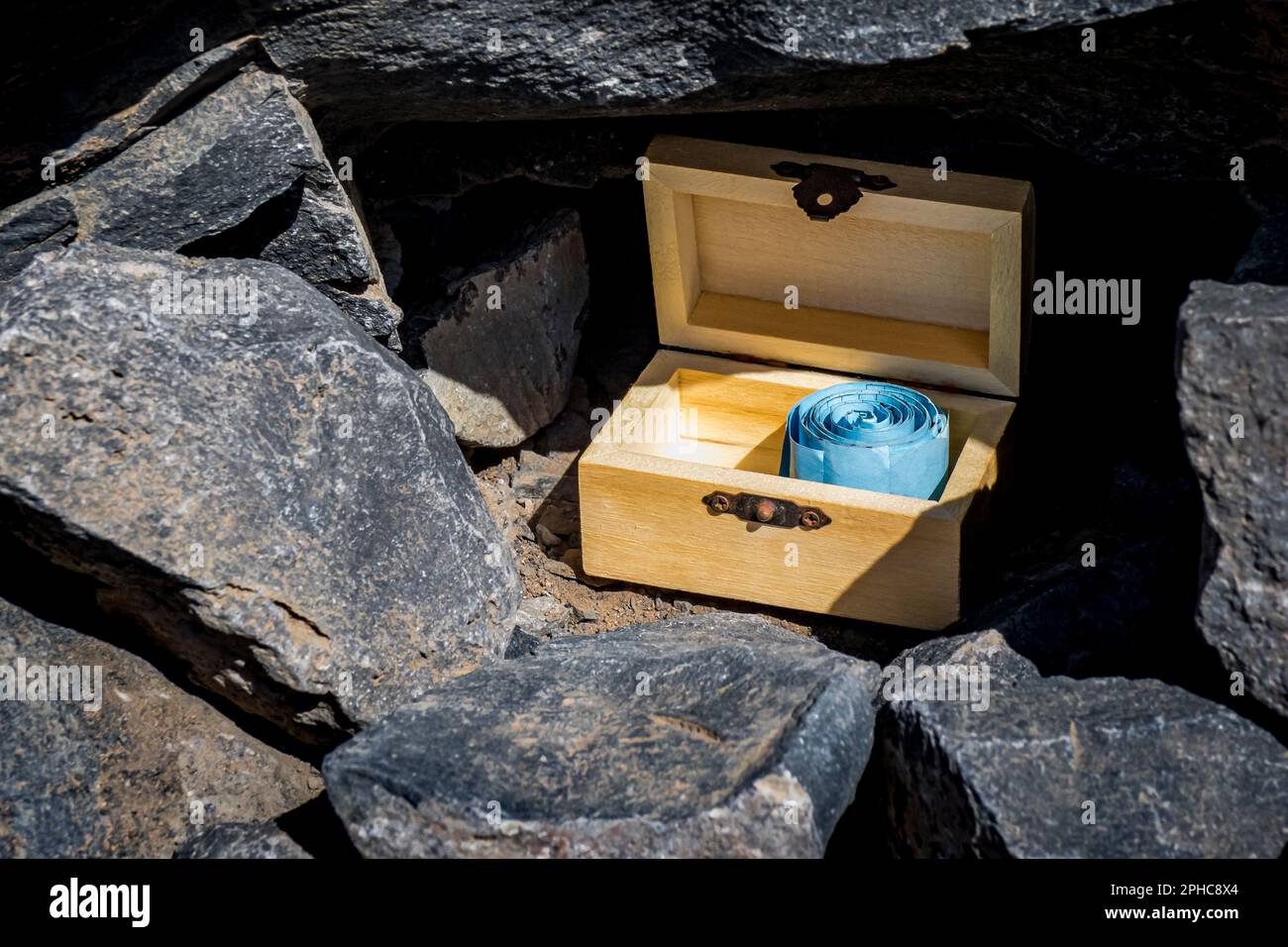 Nahaufnahme einer offenen hölzernen Geocaching-Schatztruhe mit einem blau gerollten Logbuch befindet sich versteckt in einem kleinen Loch inmitten schwarzer Steine. Stockfoto
