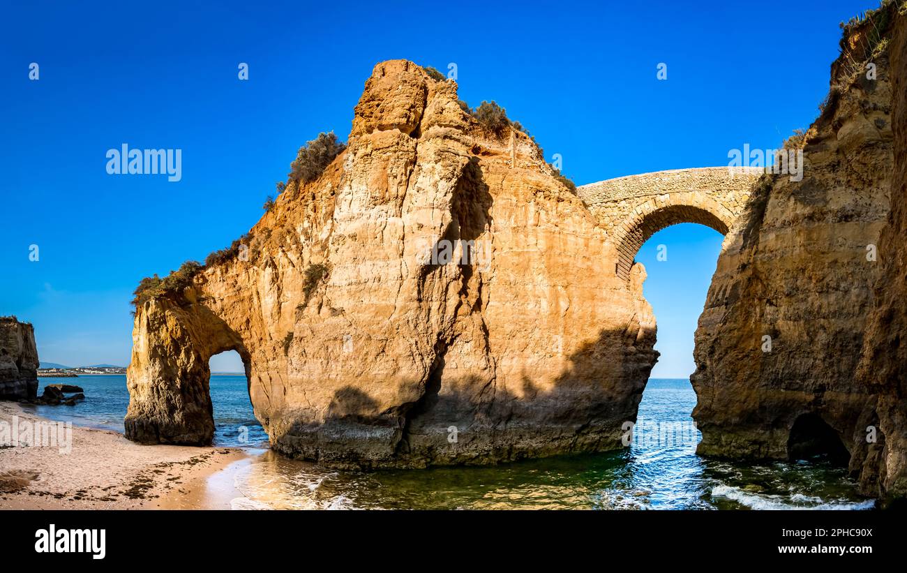 Blick aus dem niedrigen Winkel auf die Ruinen der antiken römischen Fußgängerbrücke Ponte Romana de Lagos, die zwei Klippen am Strand Praia dos Estudantes verbindet, nahe Lagos. Stockfoto