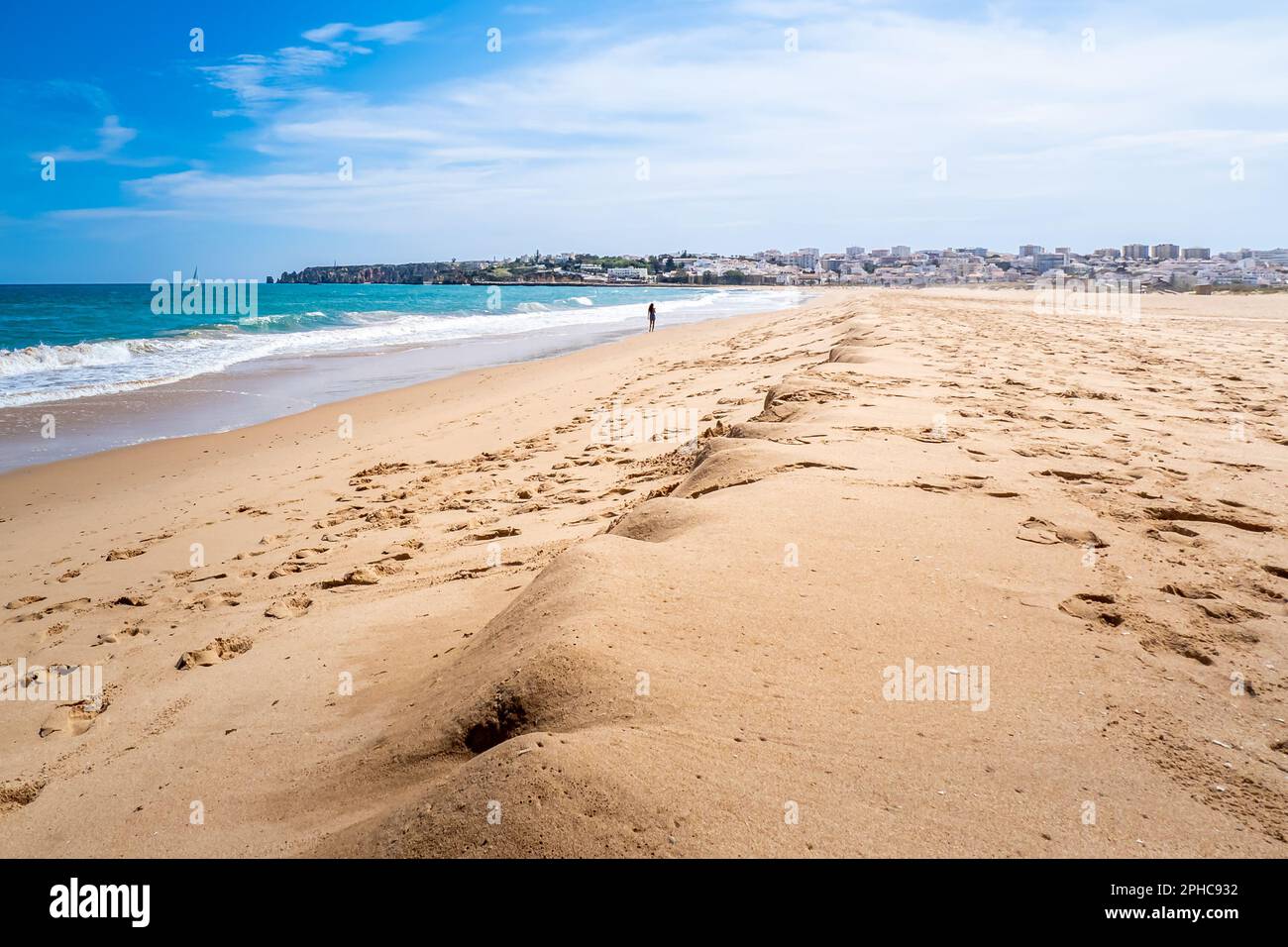 Ein atemberaubender Panoramablick auf den verlassenen Meia Praia Strand mit dem flachen Wasser, das den blauen Himmel reflektiert, und eine alleinstehende Frau, die in Richtung Lagos geht. Stockfoto