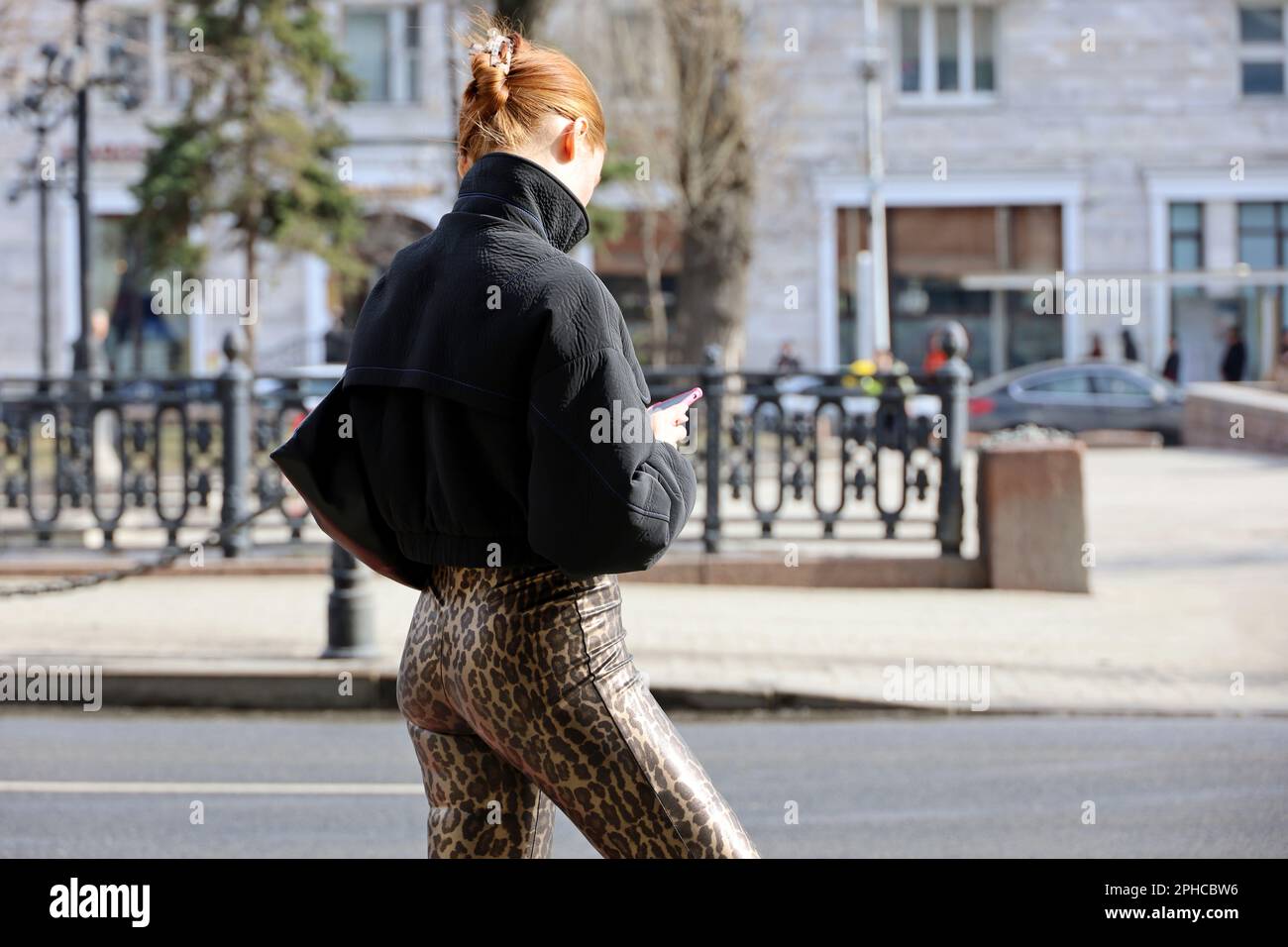 Rothaarige Frau in Hose mit Leopardenmuster, die mit einem Smartphone auf einer Straße steht. Weibliche Mode, die in der Frühlingsstadt mit dem Handy telefoniert Stockfoto