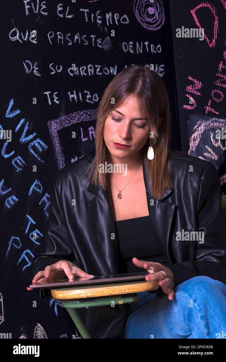 Wunderschöne junge Frau mit langen Haaren, gekleidet in einer schwarzen Lederjacke, die auf dem Schulstuhl sitzt, mit dem schwarzen Hintergrund voller geschriebener Sätze Stockfoto