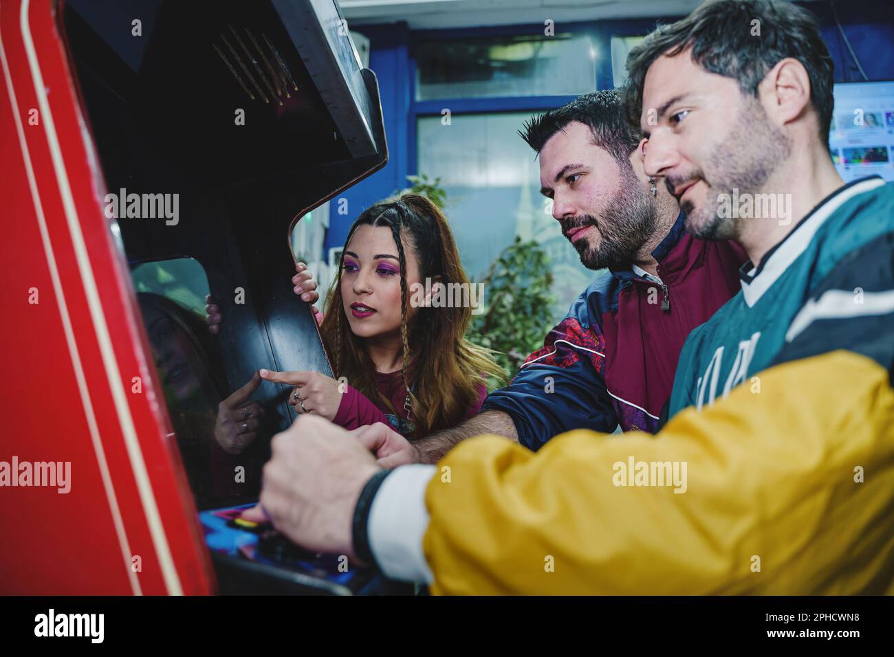 Kaukasische Freunde in 90s-Kleidung spielen ein klassisches Arkadenspiel, zwei junge Männer und eine Frau sehen sich das Spiel aufmerksam an. Stockfoto